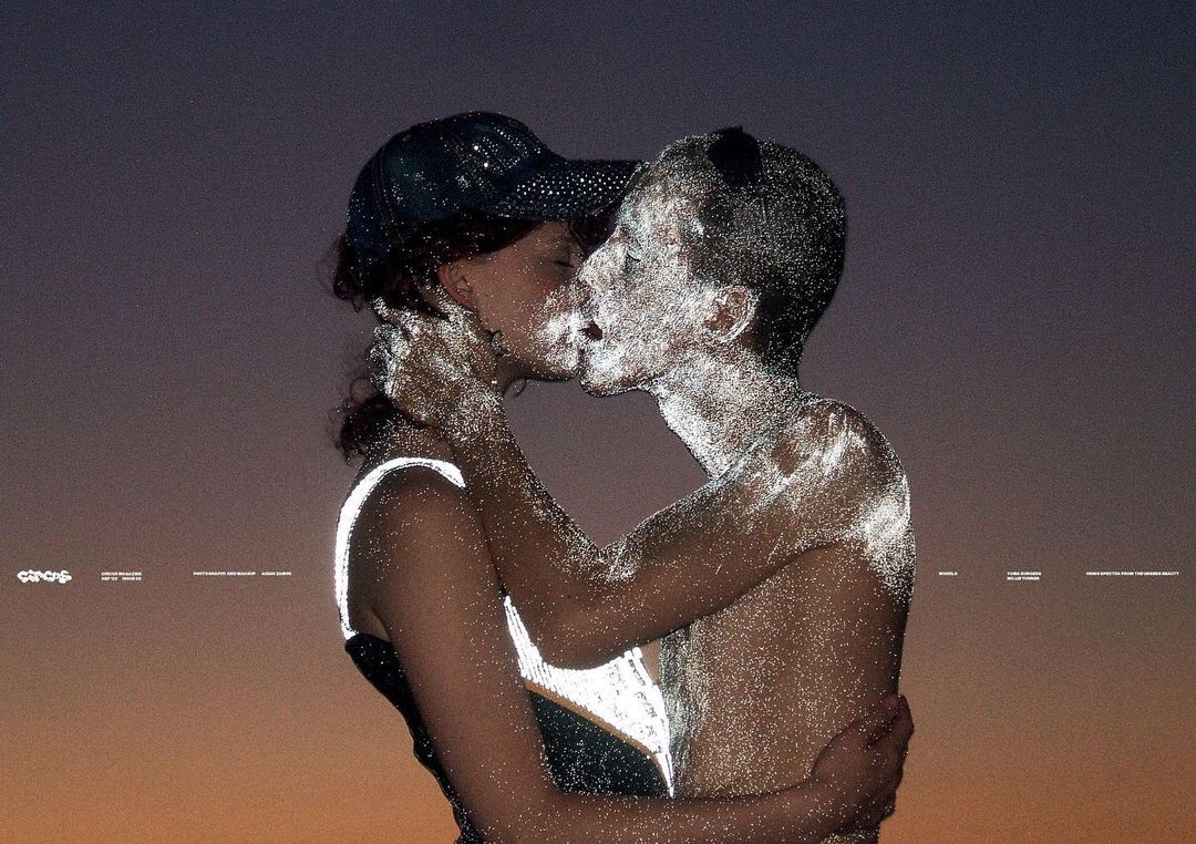 Artistic photo of a couple, heavily textured with bright dots, embracing and kissing against a gradated gray-to-orange sky at dusk.