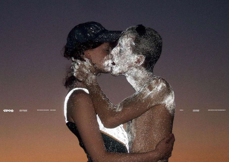 Artistic photo of a couple, heavily textured with bright dots, embracing and kissing against a gradated gray-to-orange sky at dusk.