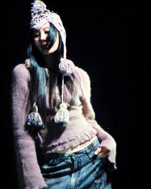 A young woman poses in a lilac sweater, jeans, and a colorful knitted hat with tassels against a dark background, looking slightly downward.