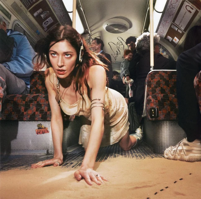 Woman crawls on sand-covered floor of subway car, wearing headphones, jewelry and dress, with other passengers in the background.
