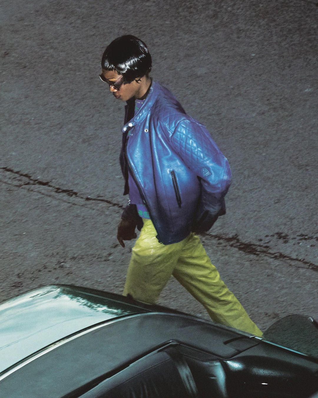 Stylish adult female wearing a blue leather jacket and yellow pants walks on an asphalt road near the hood of a dark car.