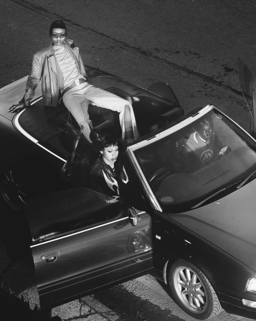 Monochrome shot of three fashion models posing in and on a convertible car with an open door on a cracked asphalt street.