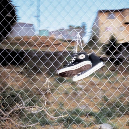 Black and white Converse high-tops hang from a wire fence outdoors against a blue sky with blurred buildings in the background.