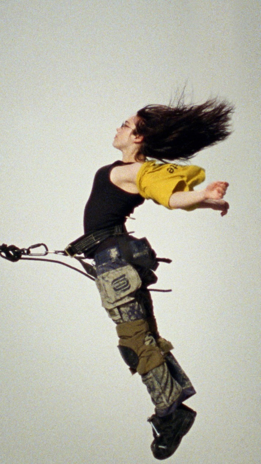 A person is suspended in mid-air wearing a black tank top, yellow shirt, patterned pants, and black shoes, with dark hair flowing upwards.