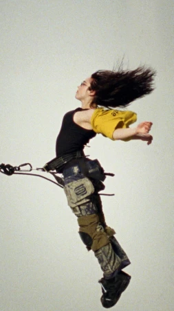A person is suspended in mid-air wearing a black tank top, yellow shirt, patterned pants, and black shoes, with dark hair flowing upwards.