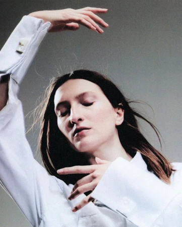 Portrait of a woman wearing a white long-sleeve shirt with arms raised in a graceful pose against a grey background.