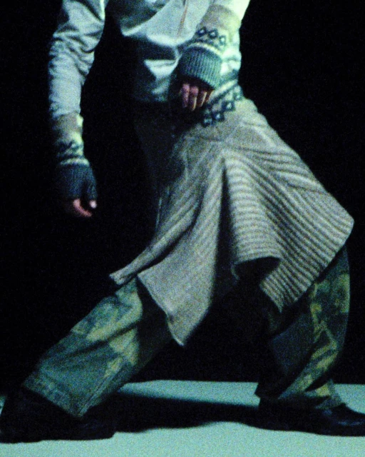Person in patterned knitwear, grey skirt, and camouflage pants strikes a dynamic pose against a dark backdrop in a dimly lit setting.