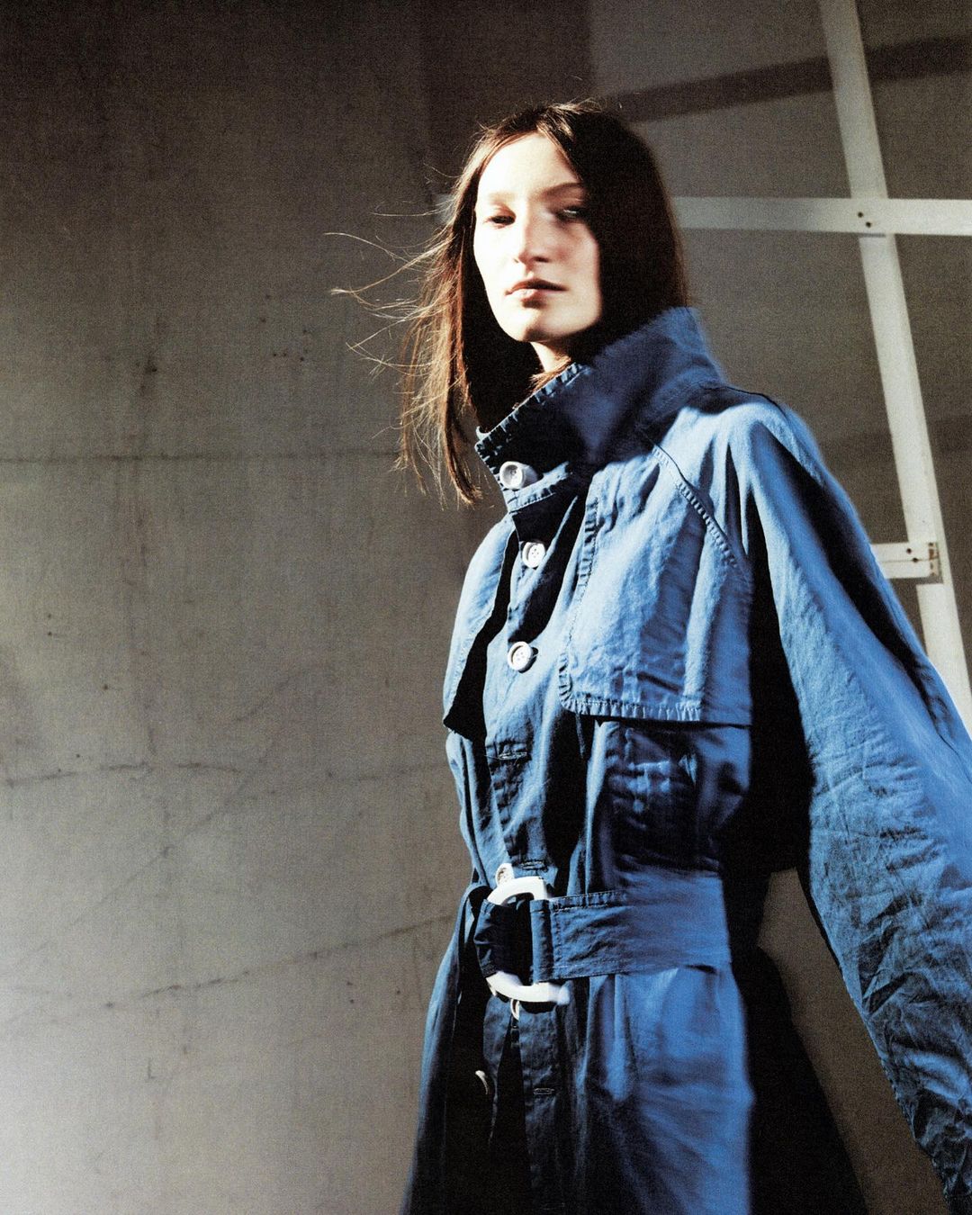 An adult female model in a blue belted trench coat poses in front of a gray concrete wall with a white ladder for a fashion editorial.