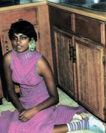 Woman in sleeveless pink patterned dress and striped socks seated on kitchen floor by wooden cabinets