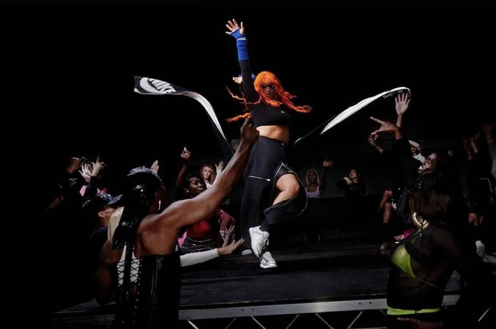 Female dancer with orange hair in a black top and black pants performs for a crowd holding a black and white flag in a dark studio.