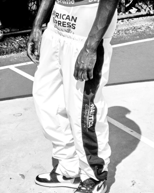 Close-up in black and white of a person wearing white athletic pants, with visible logos, and black and white sneakers on an outdoor surface.