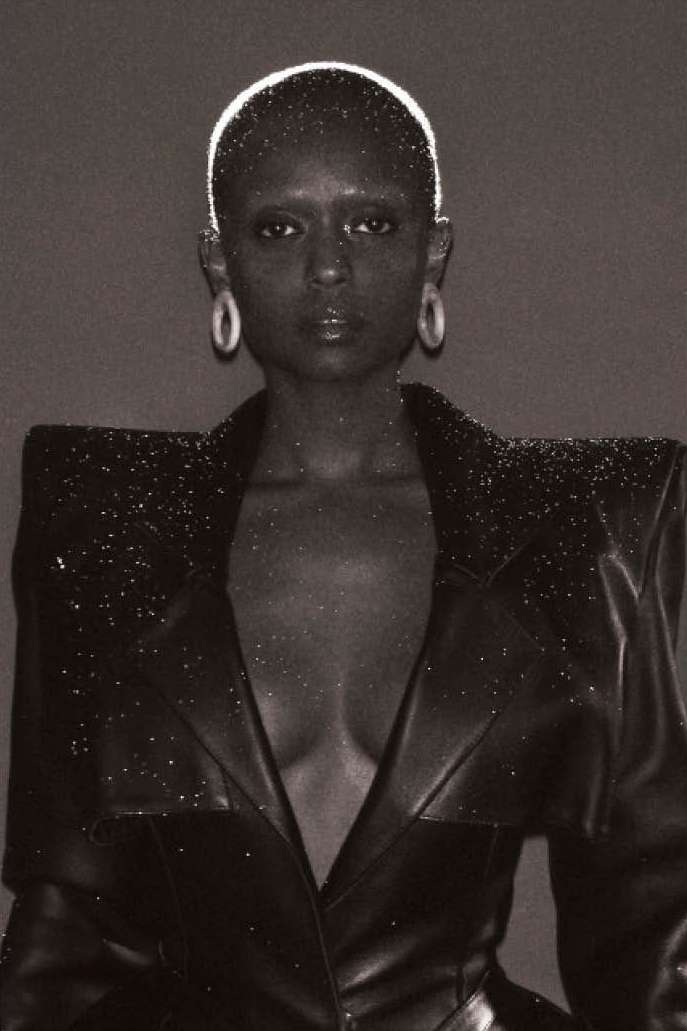 Black and white portrait of a bald Black woman wearing a studded blazer, posing against a dark grey backdrop with soft, even lighting.