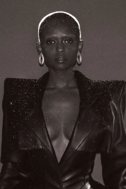 Black and white portrait of a bald Black woman wearing a studded blazer, posing against a dark grey backdrop with soft, even lighting.