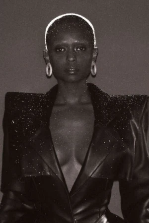 Black and white portrait of a bald Black woman wearing a studded blazer, posing against a dark grey backdrop with soft, even lighting.