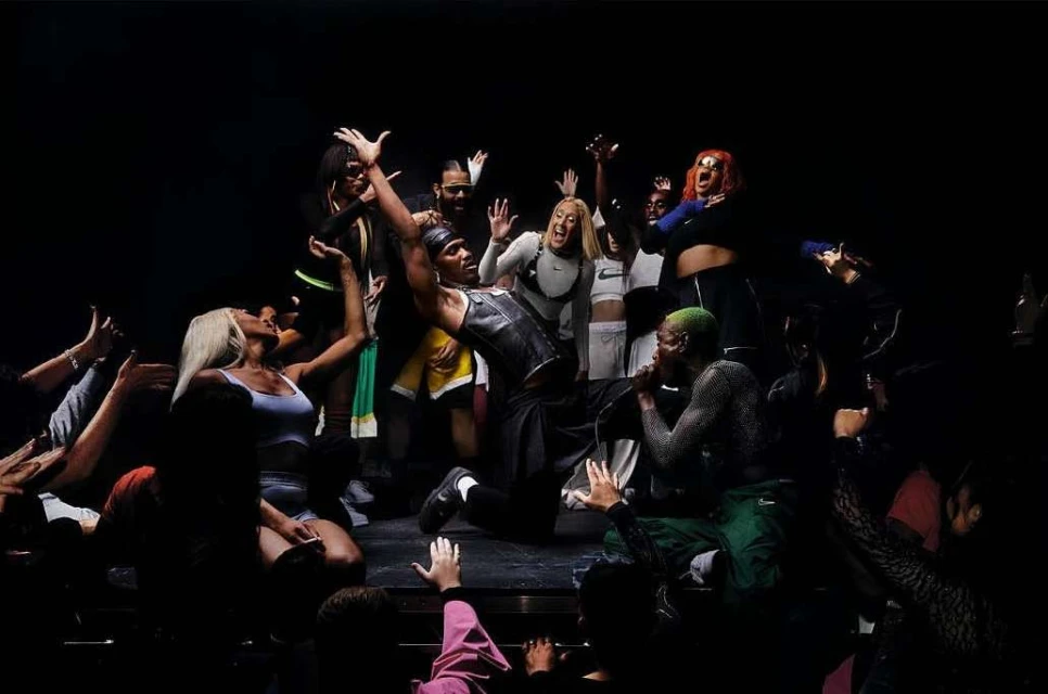 A crowd of young adults surrounds a performer on a stage, lit by stage lighting in a darkened theatre, with raised hands and excited expressions.