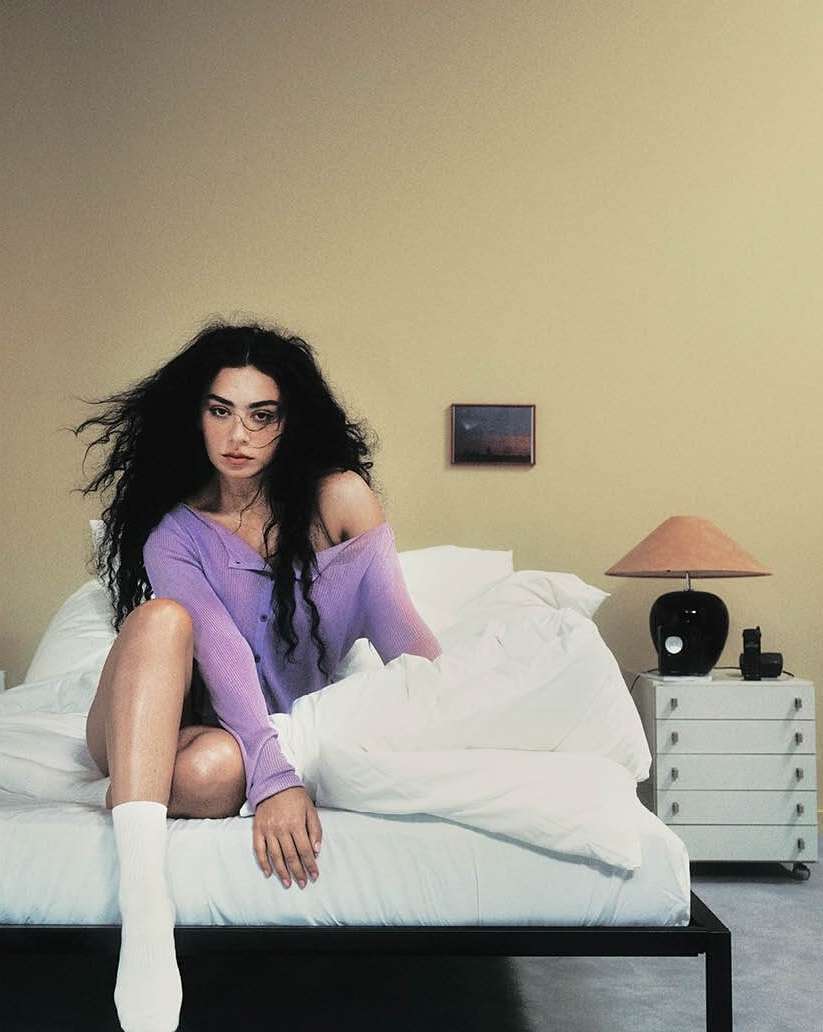 Brunette woman with long hair in a purple sweater sits on a bed with a white duvet in an indoor setting with a nightstand.