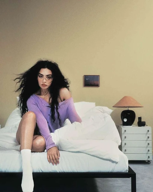 Brunette woman with long hair in a purple sweater sits on a bed with a white duvet in an indoor setting with a nightstand.