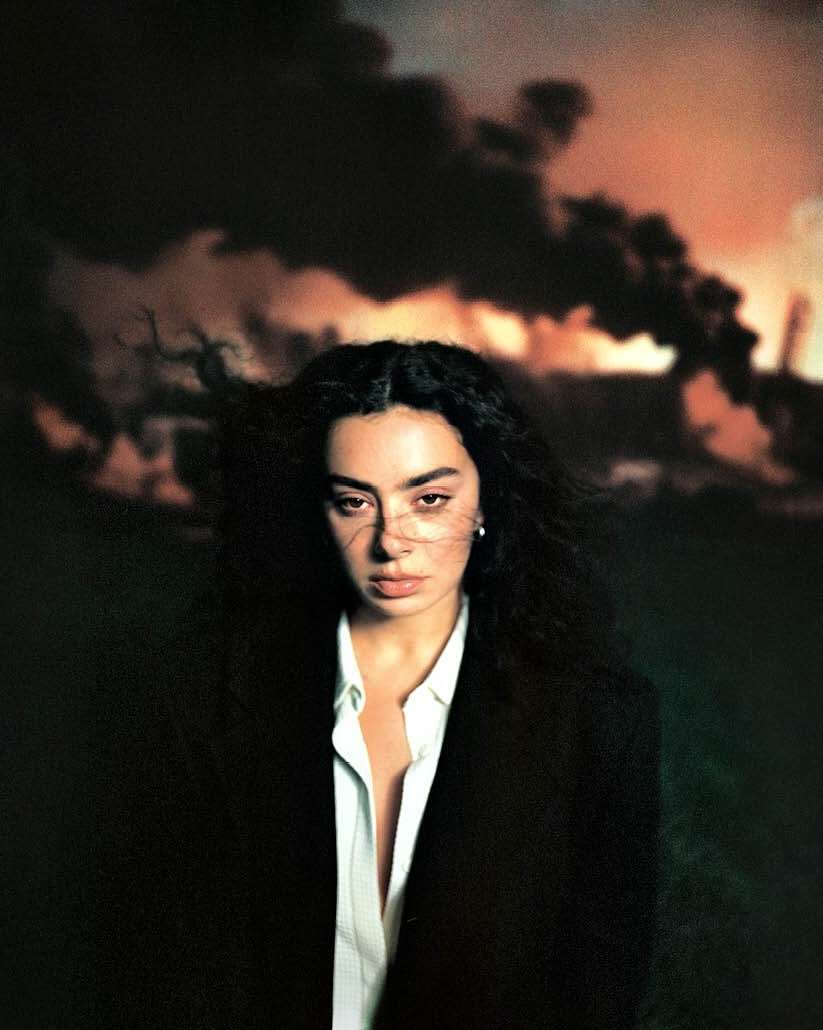 Portrait of a woman with curly hair wearing an oversized black blazer, posing in front of a dark industrial landscape at dusk.
