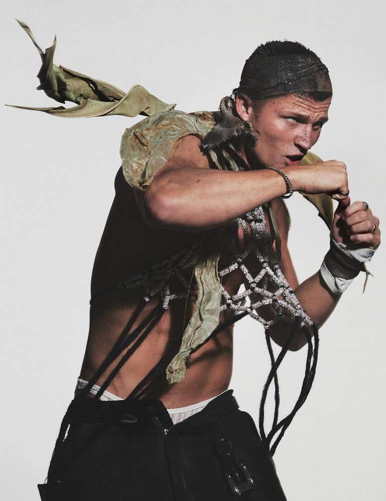 Studio shot of a man with an edgy style, wearing a net cap, deconstructed outfit, and black pants, posing in a fighting stance on a white background.