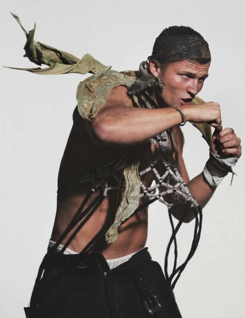 Studio shot of a man with an edgy style, wearing a net cap, deconstructed outfit, and black pants, posing in a fighting stance on a white background.