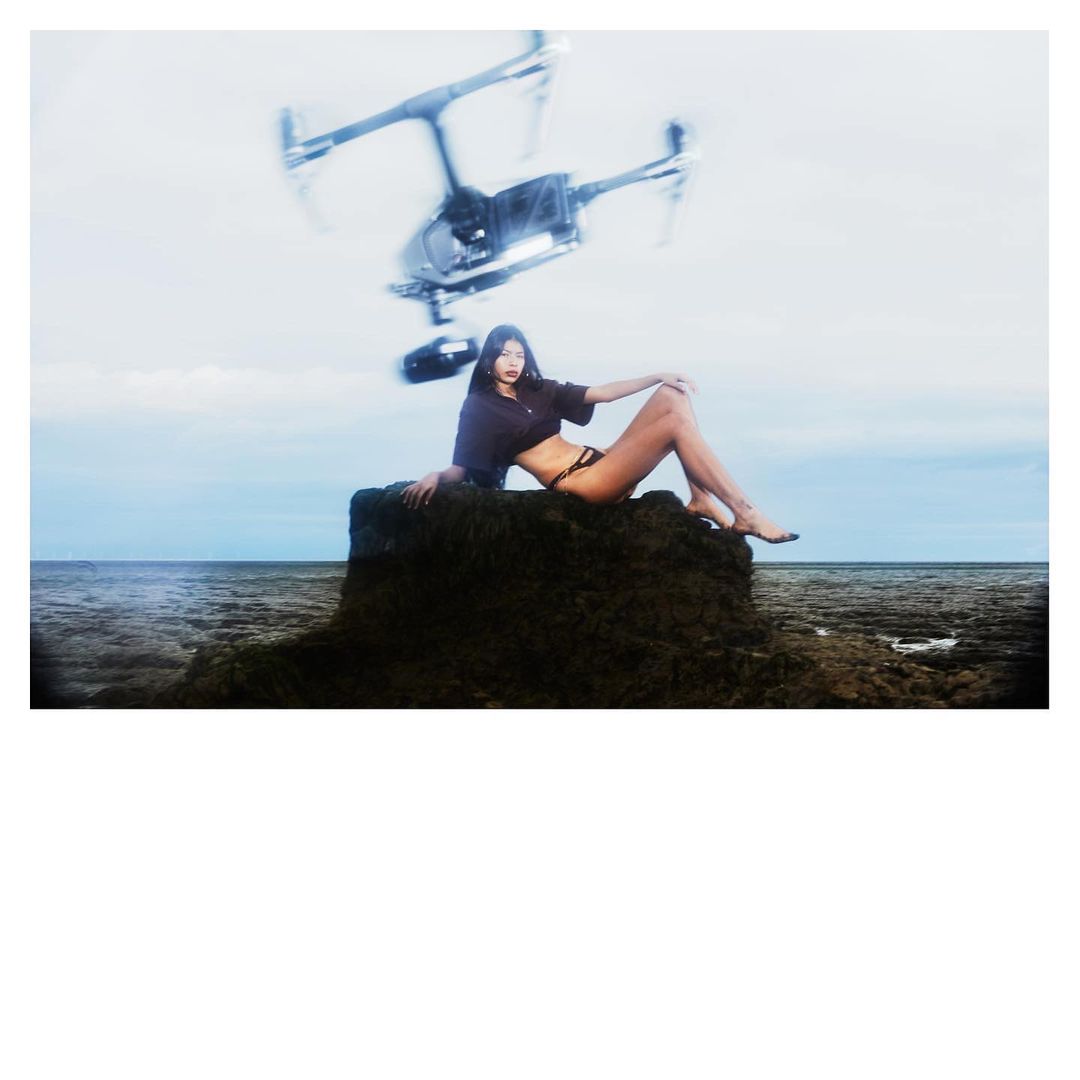 A woman sits on a rock near the ocean, with a drone hovering above her on a sunny day. Sky and water fill the background.