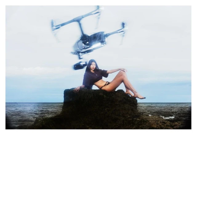 A woman sits on a rock near the ocean, with a drone hovering above her on a sunny day. Sky and water fill the background.