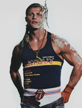 Masculine model poses indoors wearing a tank top with white and cream makeup on face and shoulder, along with a beaded necklace.