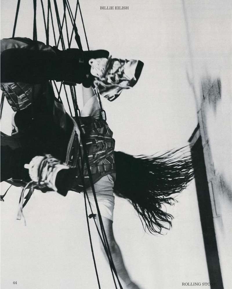 Singer Billie Eilish, suspended by ropes, in a black and white Rolling Stone magazine cover. She has long dark hair and wears a patterned vest.