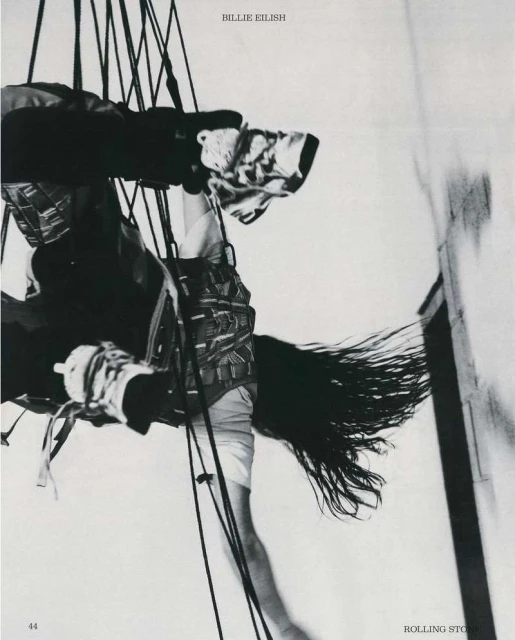 Singer Billie Eilish, suspended by ropes, in a black and white Rolling Stone magazine cover. She has long dark hair and wears a patterned vest.