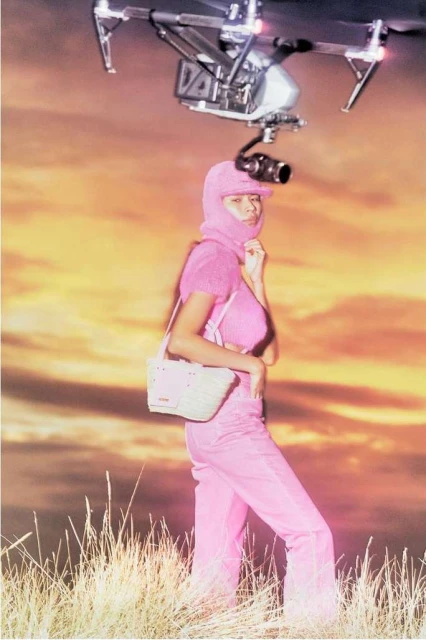 Woman in pink jumpsuit and drone with camera in a field during golden hour. Fashion shot featuring wearable technology.