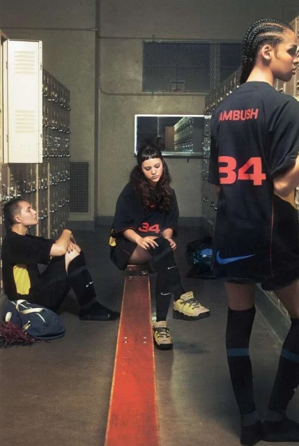 Three young adults in matching black and blue athletic apparel are seated in front of lockers for a candid fashion editorial.