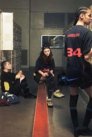Three young adults in matching black and blue athletic apparel are seated in front of lockers for a candid fashion editorial.