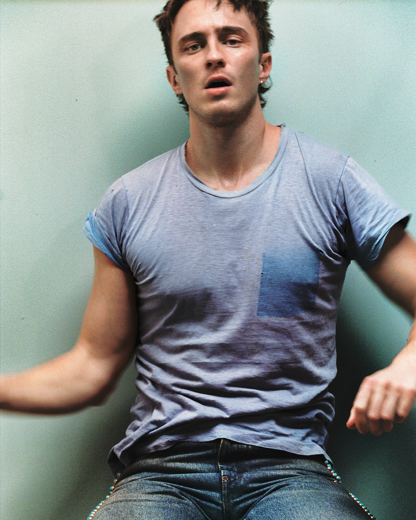 Portrait of adult male with short dark hair wearing a grey t-shirt and blue jeans indoors with soft lighting against a pale blue wall.
