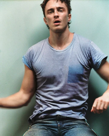Portrait of adult male with short dark hair wearing a grey t-shirt and blue jeans indoors with soft lighting against a pale blue wall.