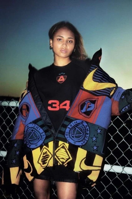 Woman in streetwear fashion wearing Nike logo patterned jacket and black top with the number 34, posing in front of chain link fence.