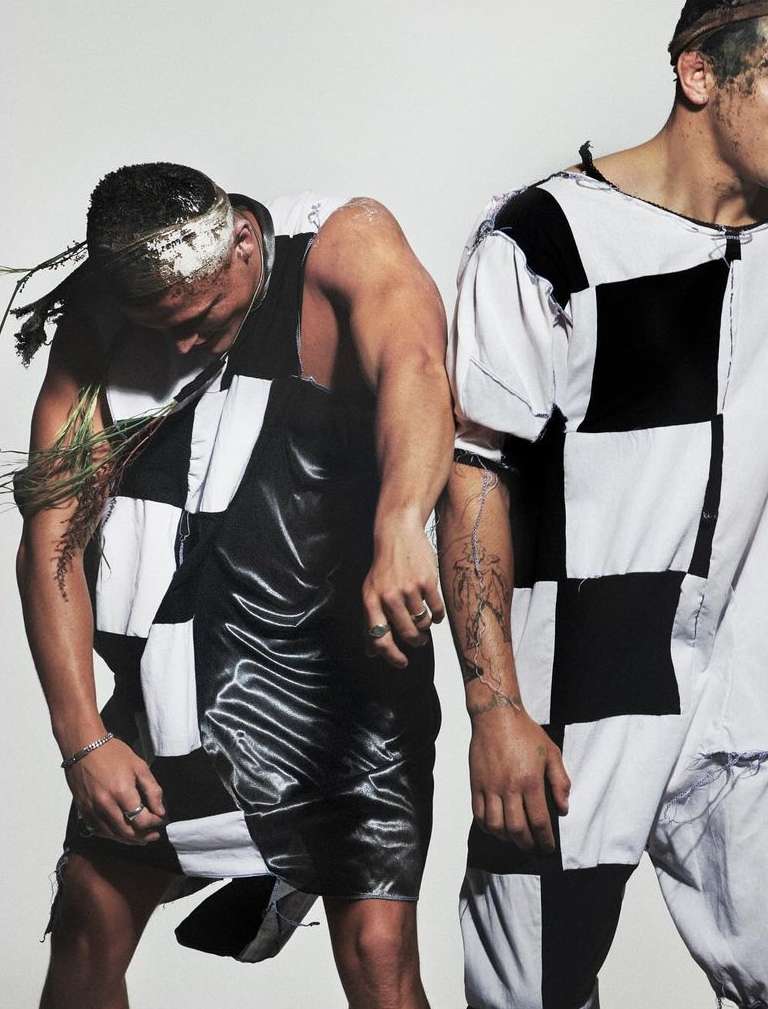 Two men wearing black and white checkered outfits standing against a plain white background by photographer Aidan Zamiri.