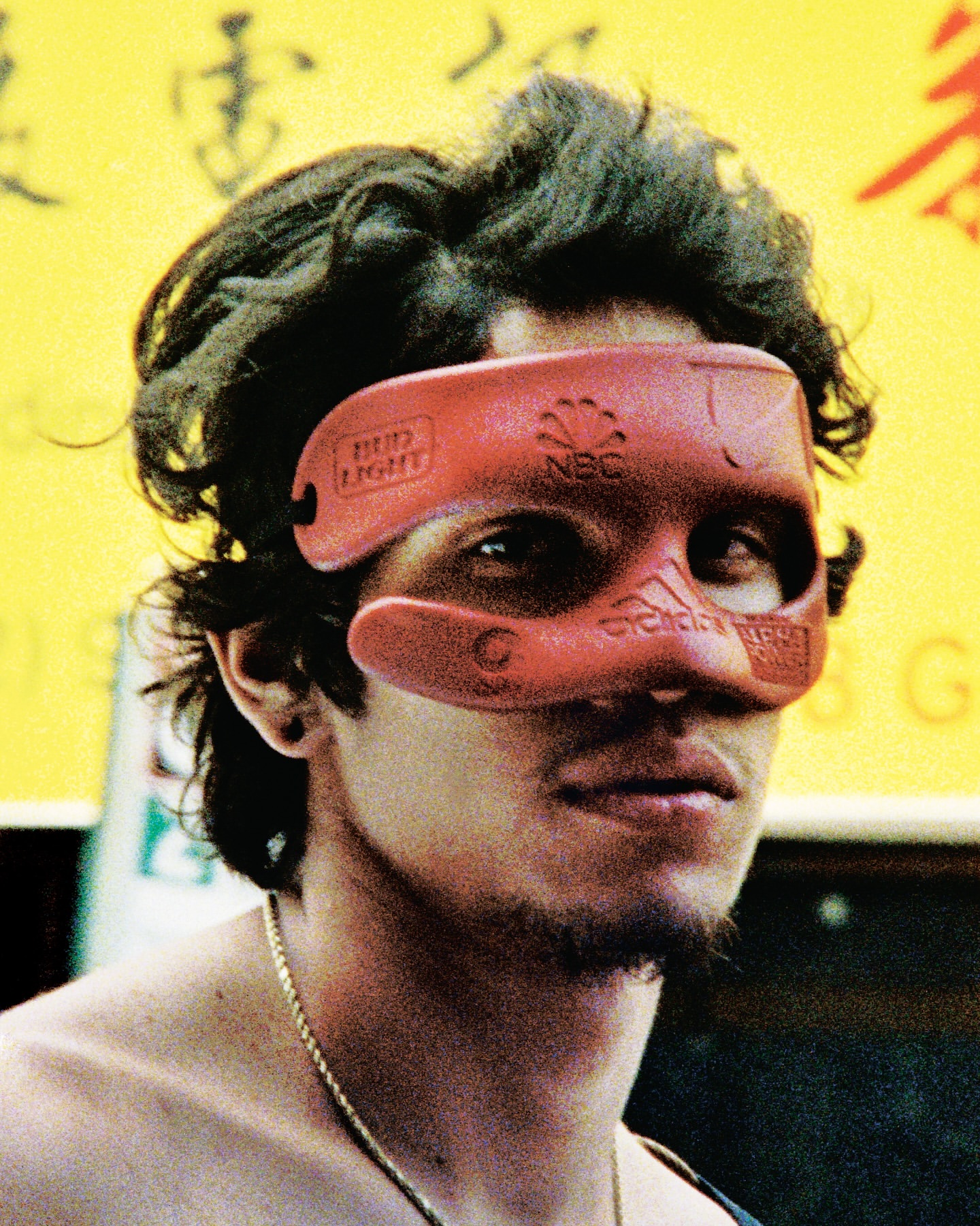 Portrait of a person wearing a unique novelty mask with logos against a blurred yellow background, shot with natural outdoor lighting.