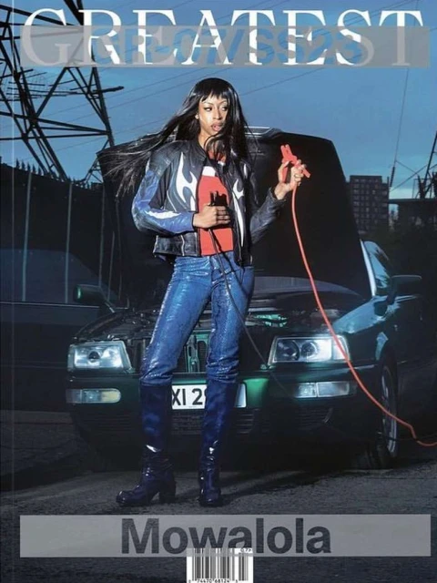 A woman with long dark hair poses with jumper cables in front of a car with its hood up, an urban scene with electrical towers.