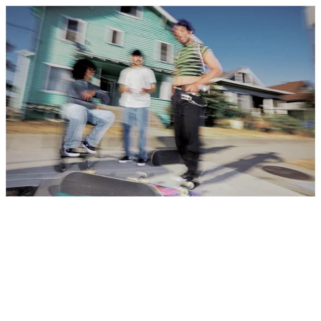 Three young adults gather on a sidewalk, skateboards nearby, in front of a light-green two-story house on a sunny day. Blurred motion effect.
