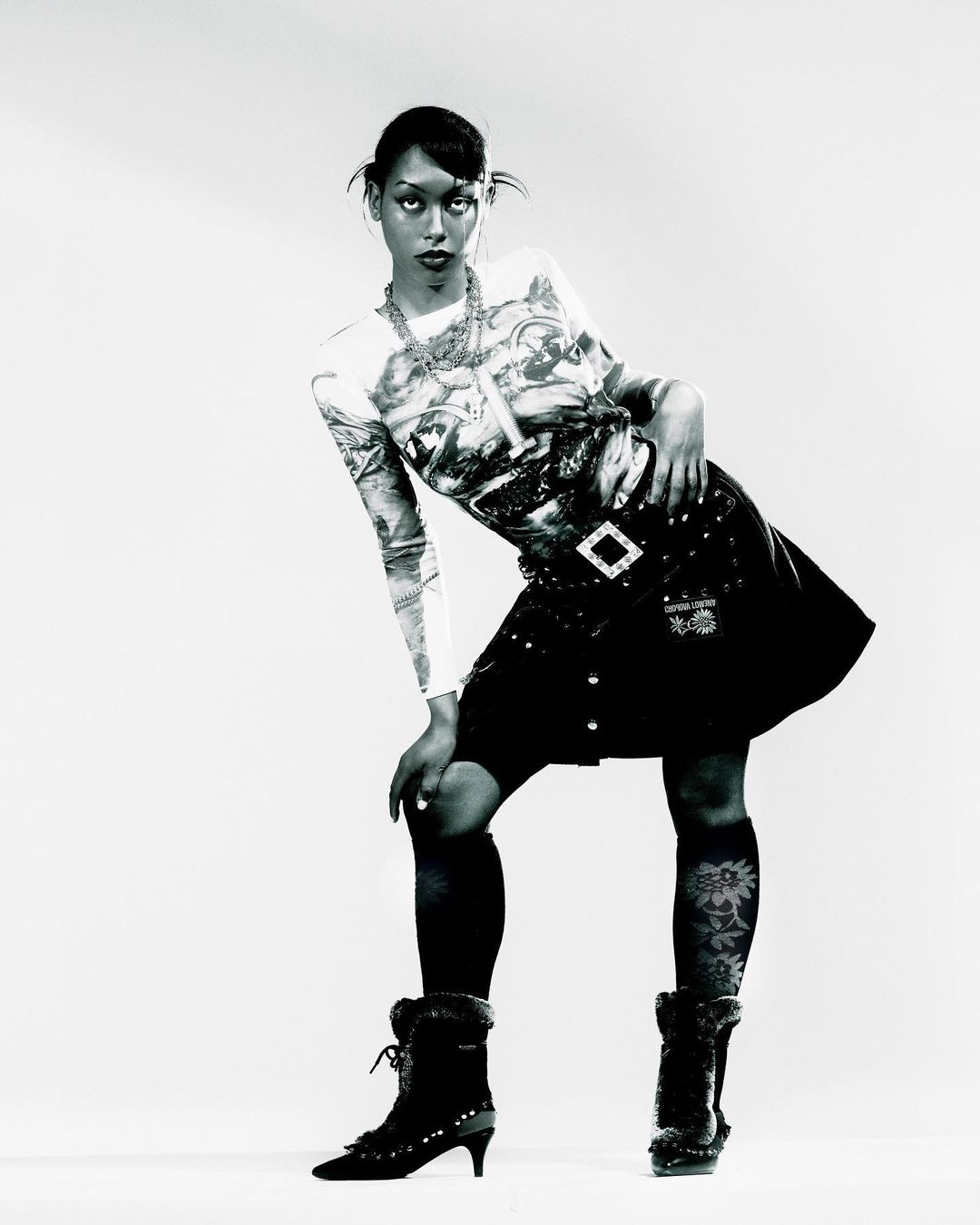 Black and white portrait of a woman posing in a patterned top and dark skirt against a plain studio background.