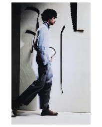 Profile of a young man with curly hair wearing a button-down shirt and trousers against a wall hung with hand tools.