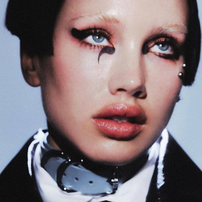 Close-up portrait of a woman with bleached eyebrows, dramatic black eyeliner, a light blue collar, and dark outerwear.