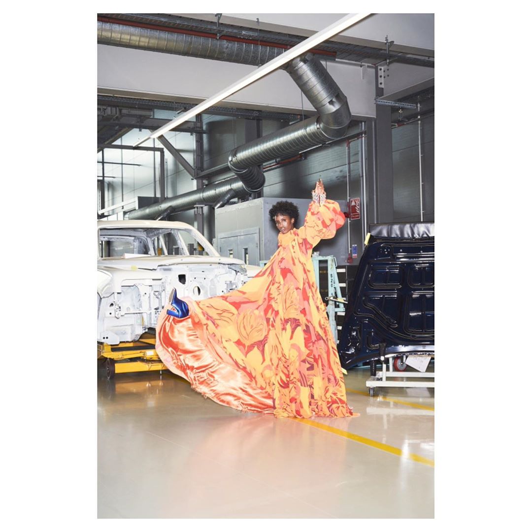 Adult female in orange gown posing in an automotive assembly facility next to a disassembled car and machinery, with overhead lighting.