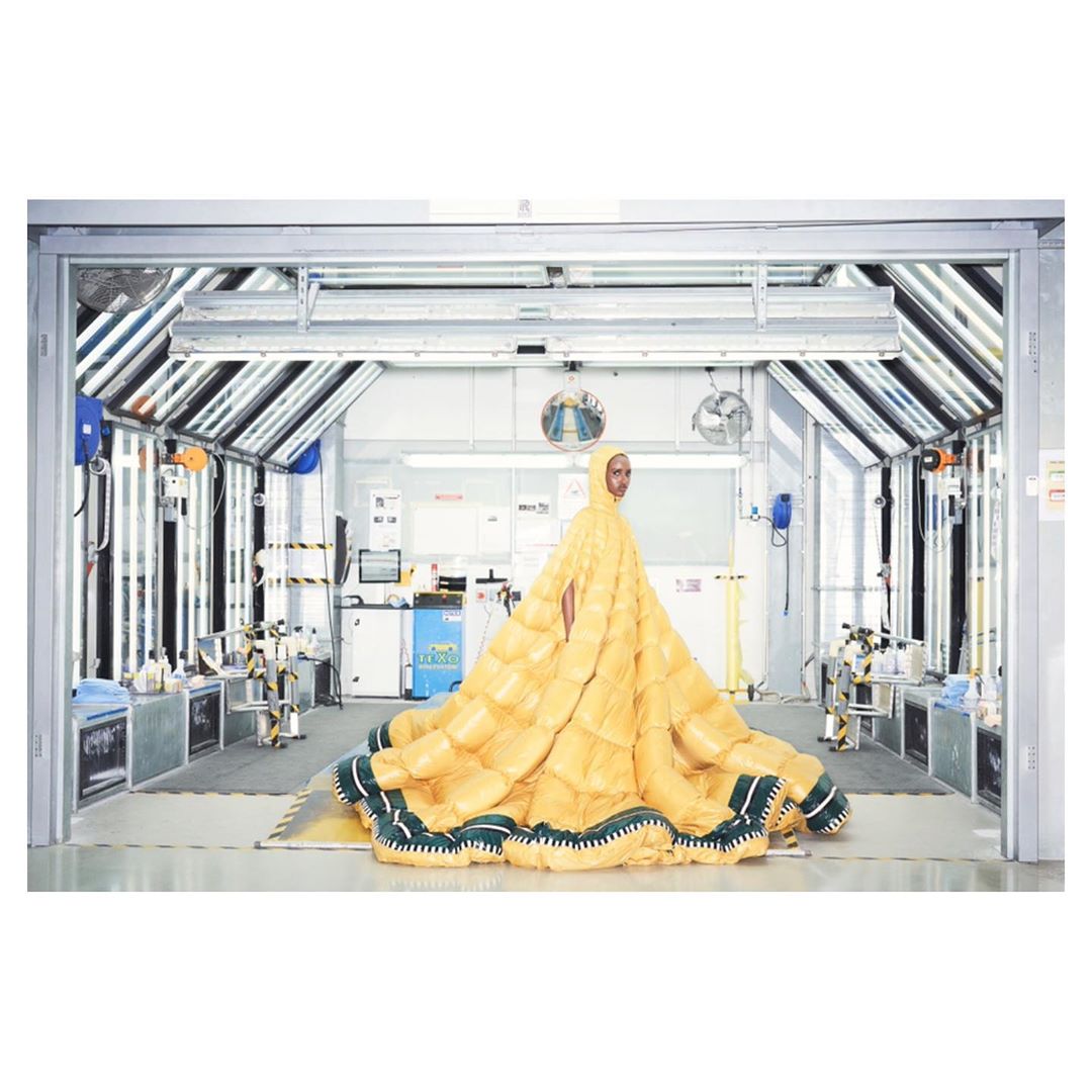 A woman poses in a yellow, quilted, hooded gown with green trim in a bright industrial setting with white walls and overhead lighting.