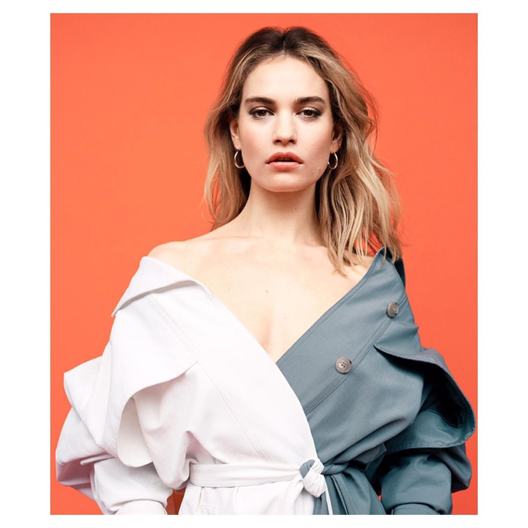Portrait of a young woman with blonde hair wearing a color-blocked trench coat against a vibrant orange backdrop, gazing directly at the viewer.