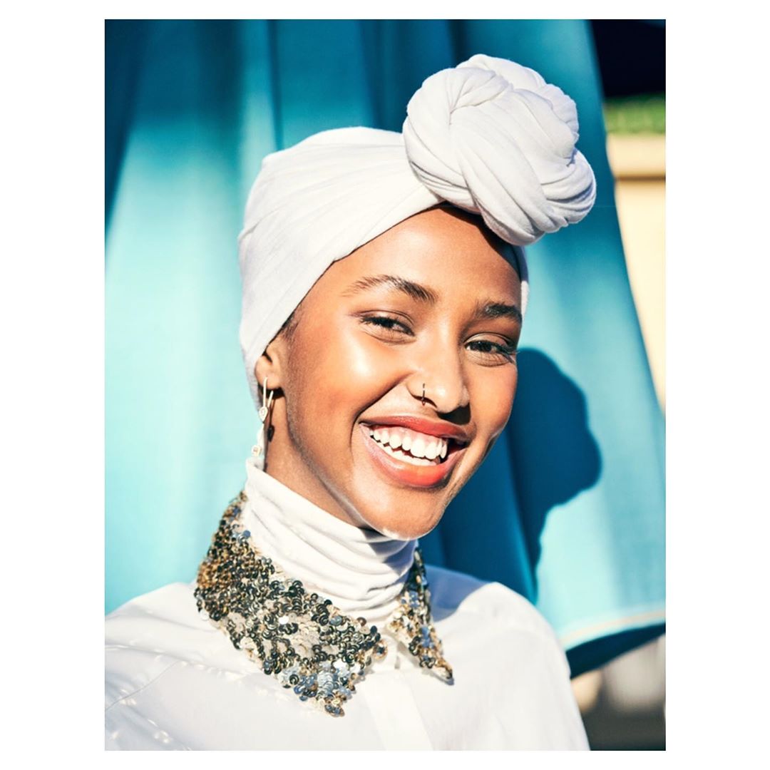 Close-up portrait of a smiling adult woman with a white headscarf and beaded collar against a blue and green background outdoors.