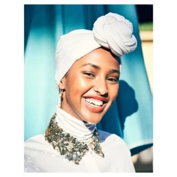Portrait of a smiling woman wearing a white turban and a sequin-collared shirt against a blue backdrop.