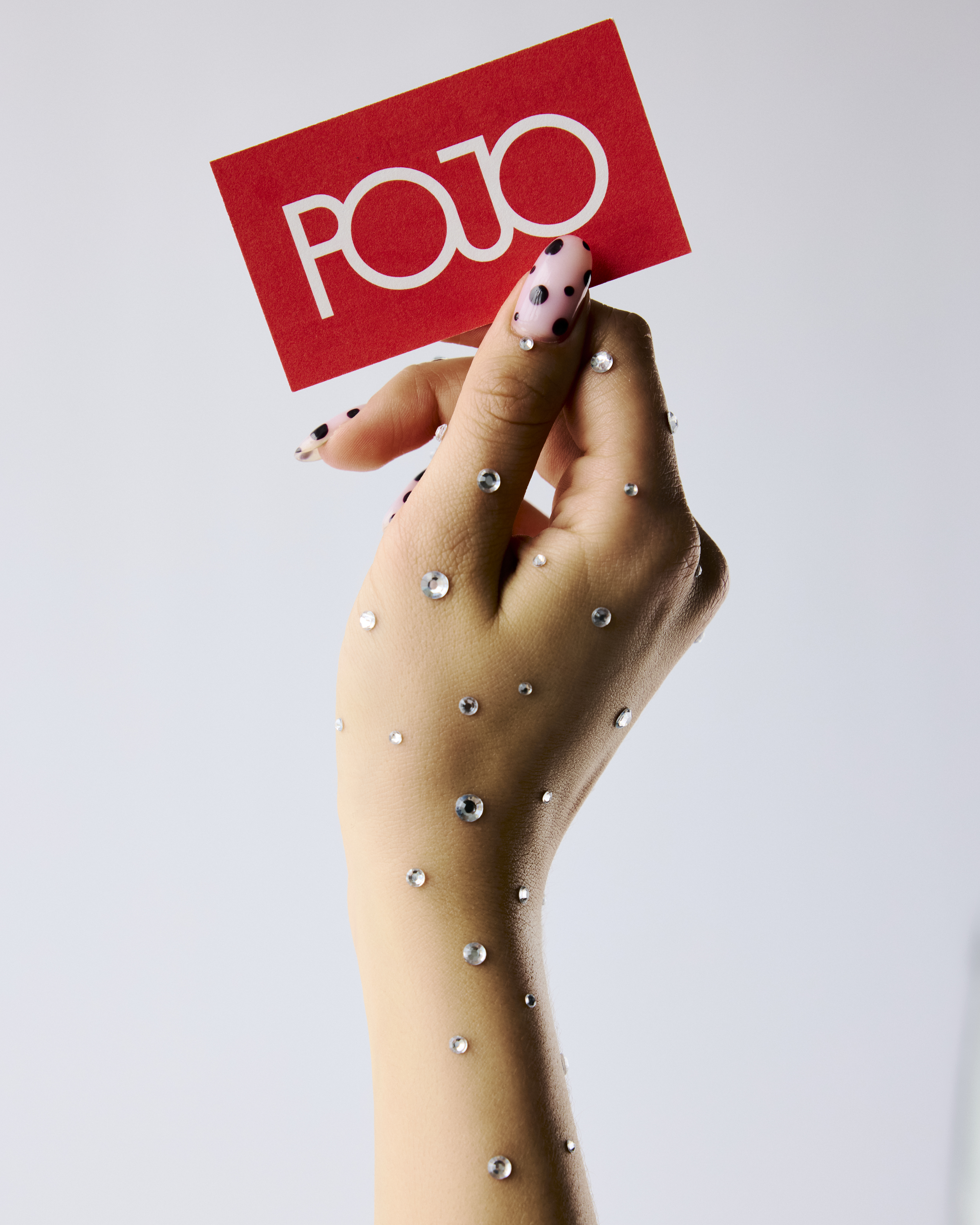 Close-up of a hand adorned with rhinestones holding a red card with white text, against a light gray background, promoting beauty.