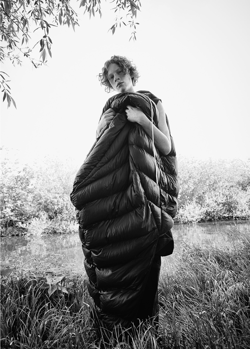 Monochrome portrait of a young person enveloped in a black puffer sleeping bag, standing in a grassy field near a body of water.