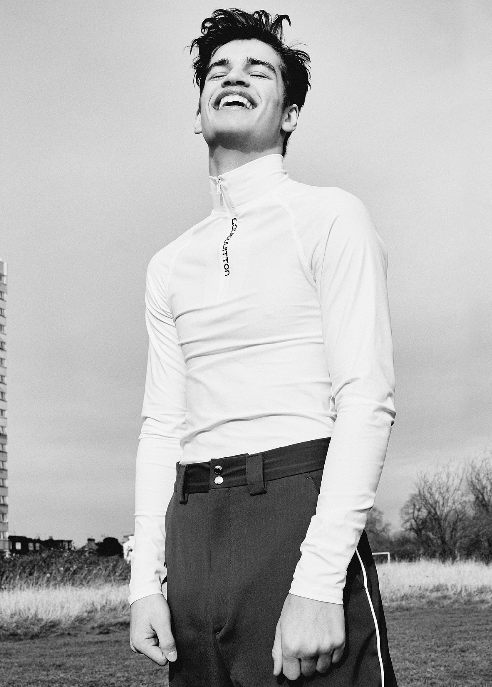 A young adult male in a white long-sleeve shirt and dark pants smiles outdoors with a building and sky in the background in a black and white image.